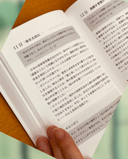 手で押さえながら開かれた書籍『一日一歩』の本文ページ。11日「休を大切に」と12日「感謝を習慣に」の見出しが見え、信仰に関する黙想文が縦に配置されている。