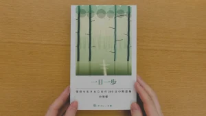 木目の机の上で、両手が一冊の本を持っている。表紙には、淡い緑色の森を思わせるイラストが描かれ、タイトルは「一日一歩」。下部に「信仰を生きるための365日の黙想集」「村思郁」「エイレーネ書店」と記載されている。